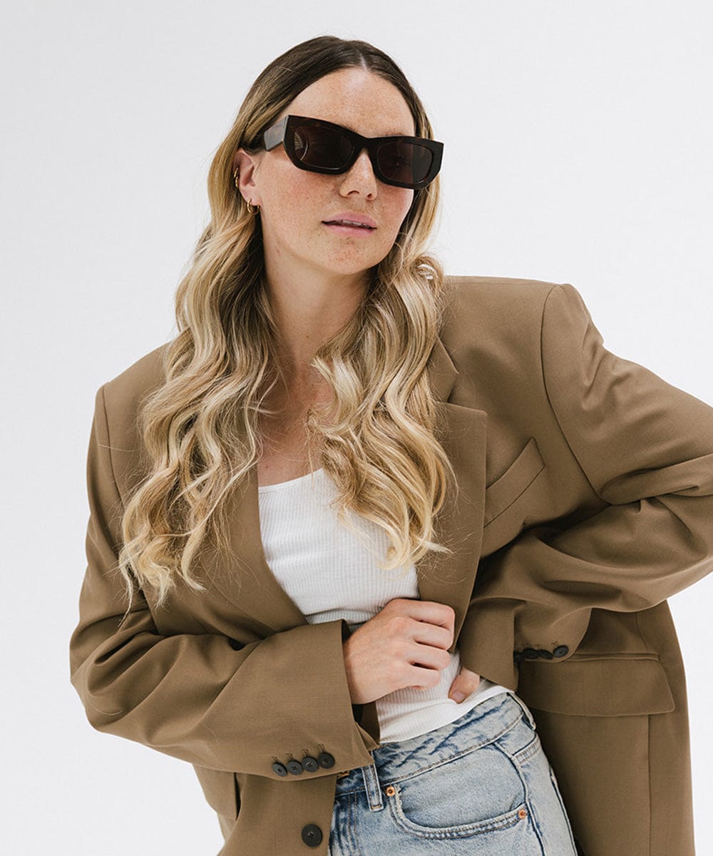 Woman wearing tortoise pattern sunglasses and a white top with a brown blazer against a plain background #color_tortoise