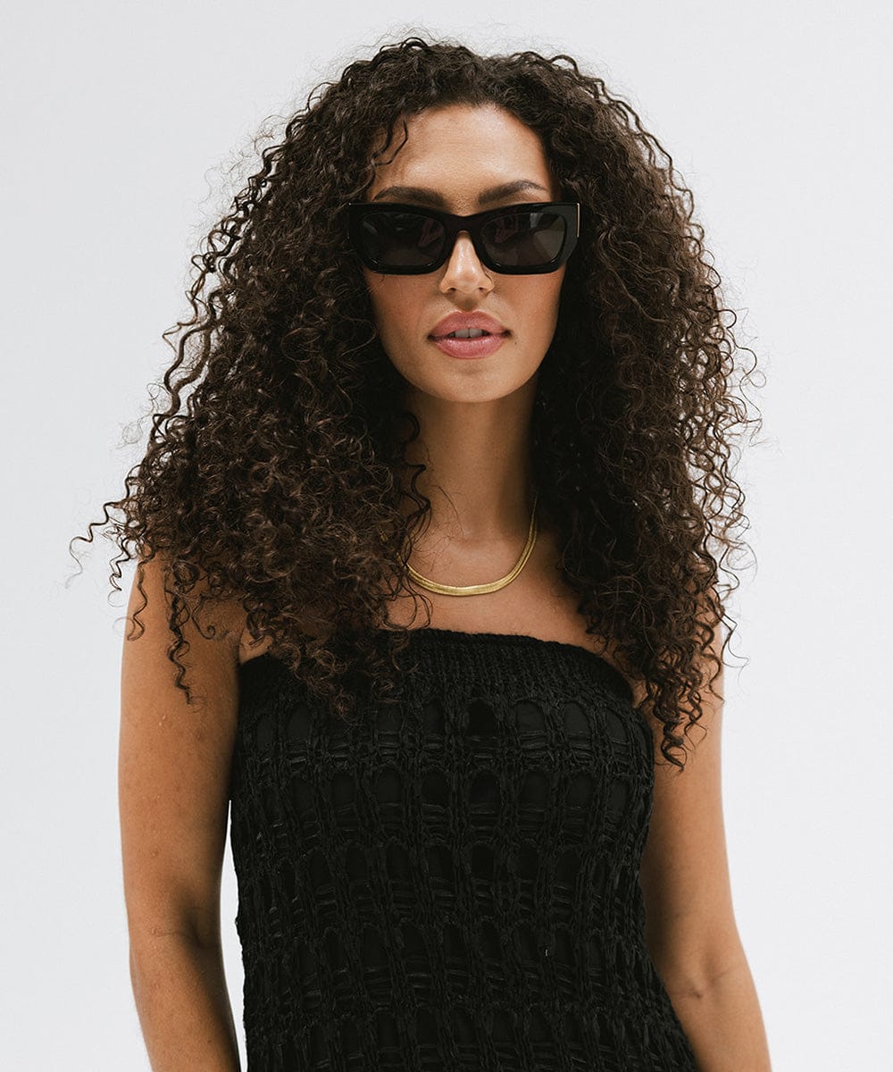 Woman wearing black sunglasses, a gold chain and a black top against a plain background #color_black