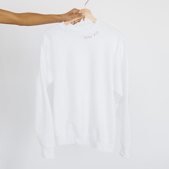 Person carrying a white sweatshirt with the text 'GIGI PIP' in beige against a plain background #color_white-beige