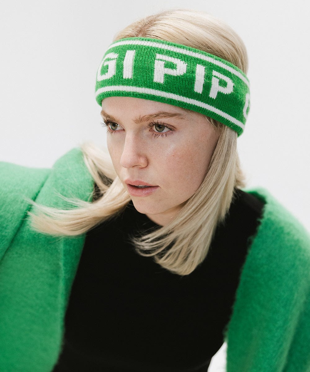 Woman wearing an evergreen colored headband with 'GIGI PIP' text and a black t-shirt with a green oversized scarf against a plain background #color_evergreen