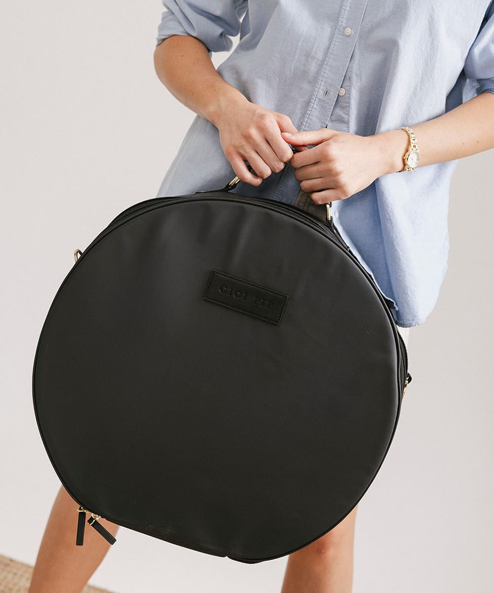 Person wearing a light blue shirt carrying a black round hat travel case against a plain background #color_black