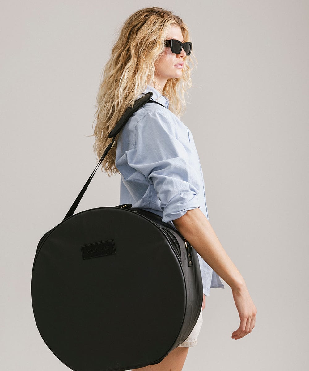 Woman wearing sunglasses and a light blue shirt carrying a black round hat travel case against a plain background #color_black