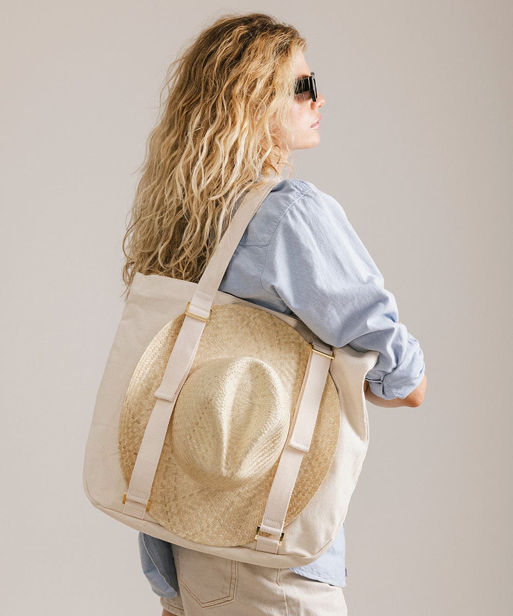 Woman wearing sunglasses, a blue shirt and white shorts, carrying a hat on a natural colored canvas tote bag, facing sideways against a plain background #color_natural