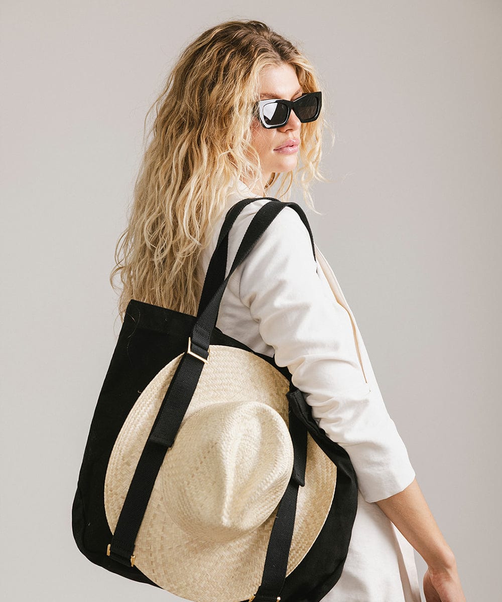 Woman wearing sunglasses and a white jacket, carrying a hat on a black canvas tote bag against a plain background #color_black