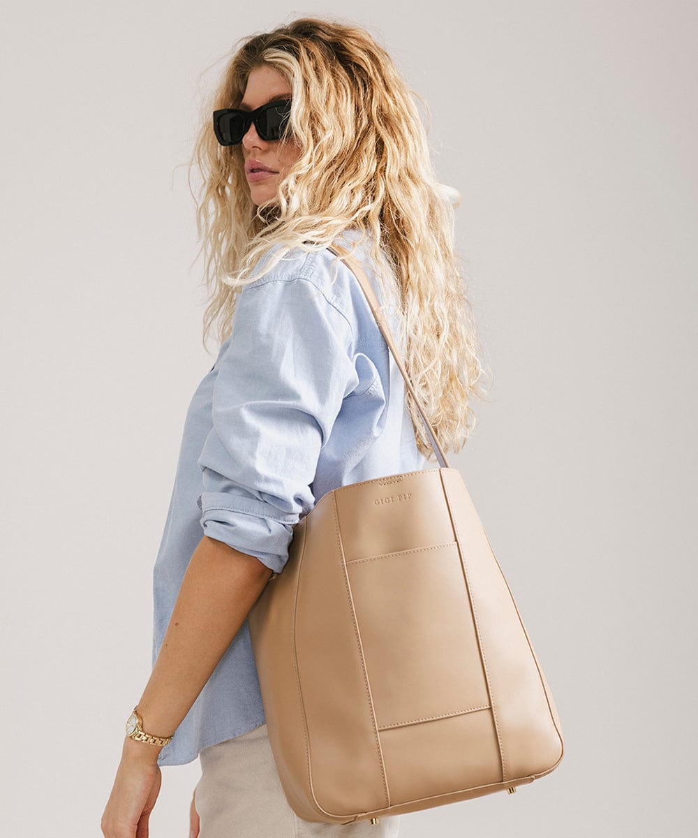 Woman wearing sunglasses, a blue shirt and cream shorts, carrying a tan Dia everyday leather tote bag, posing sideways against a plain background #color_tan