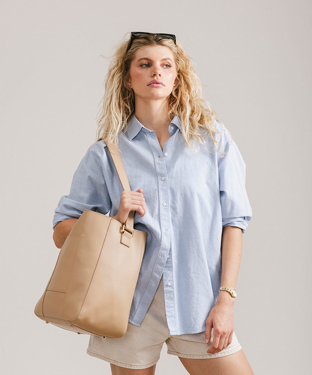 Woman wearing a blue shirt and cream shorts carrying a tan Dia everyday leather tote bag against a plain background #color_tan