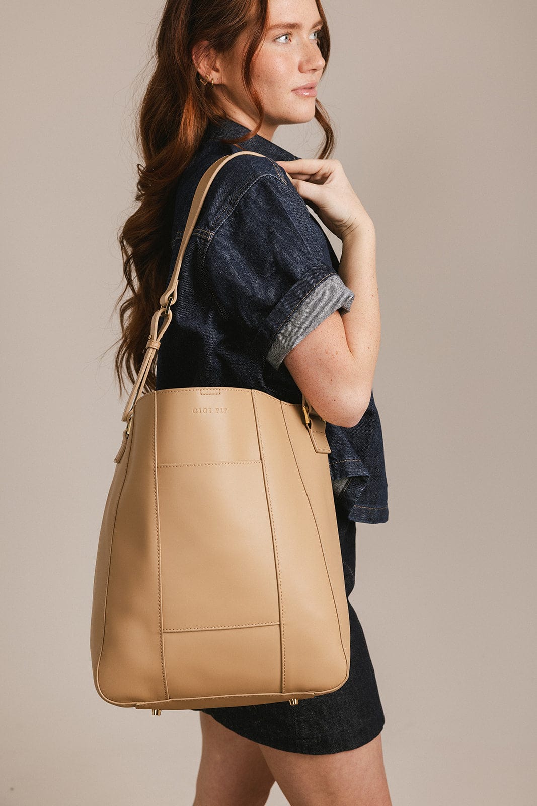 Woman wearing a denim shirt and denim skirt carrying a tan Dia everyday leather tote bag, posing sideways against a plain background #color_tan