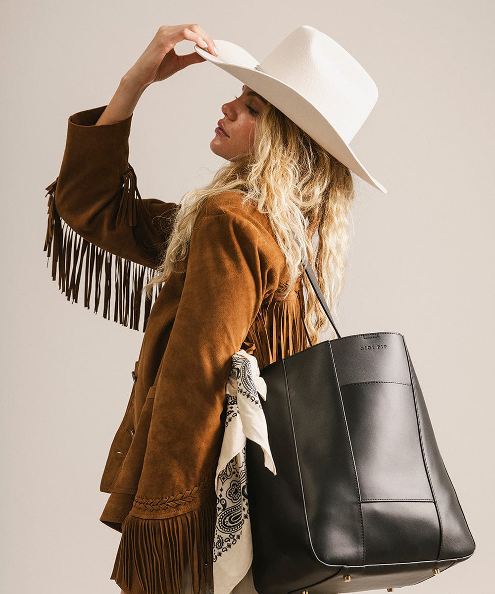 Woman wearing a brown suede jacket with fringe, white hat, and black handbag on a plain background #color_black