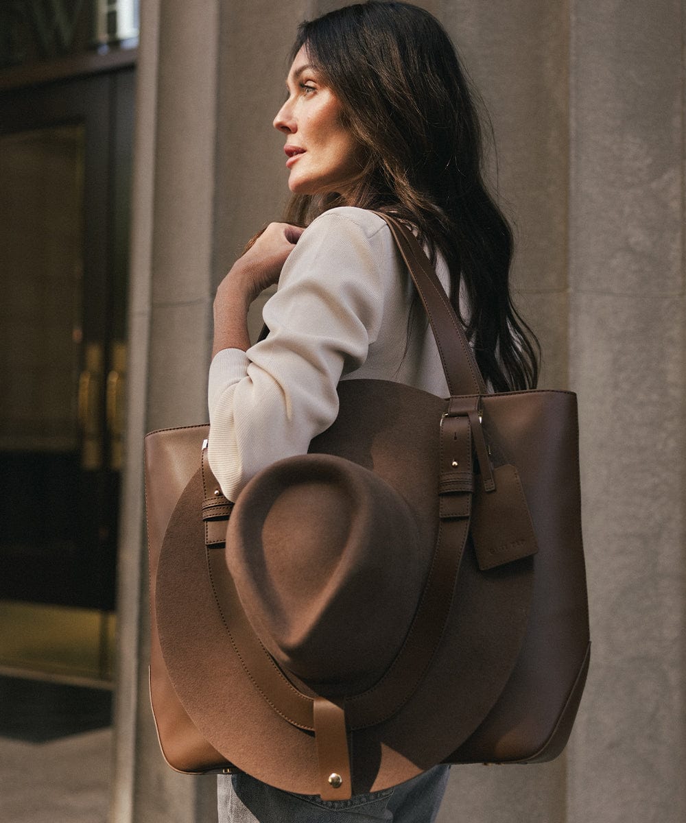 Woman holding a large brown leather bag with a hat on a city street. #color_chocolate