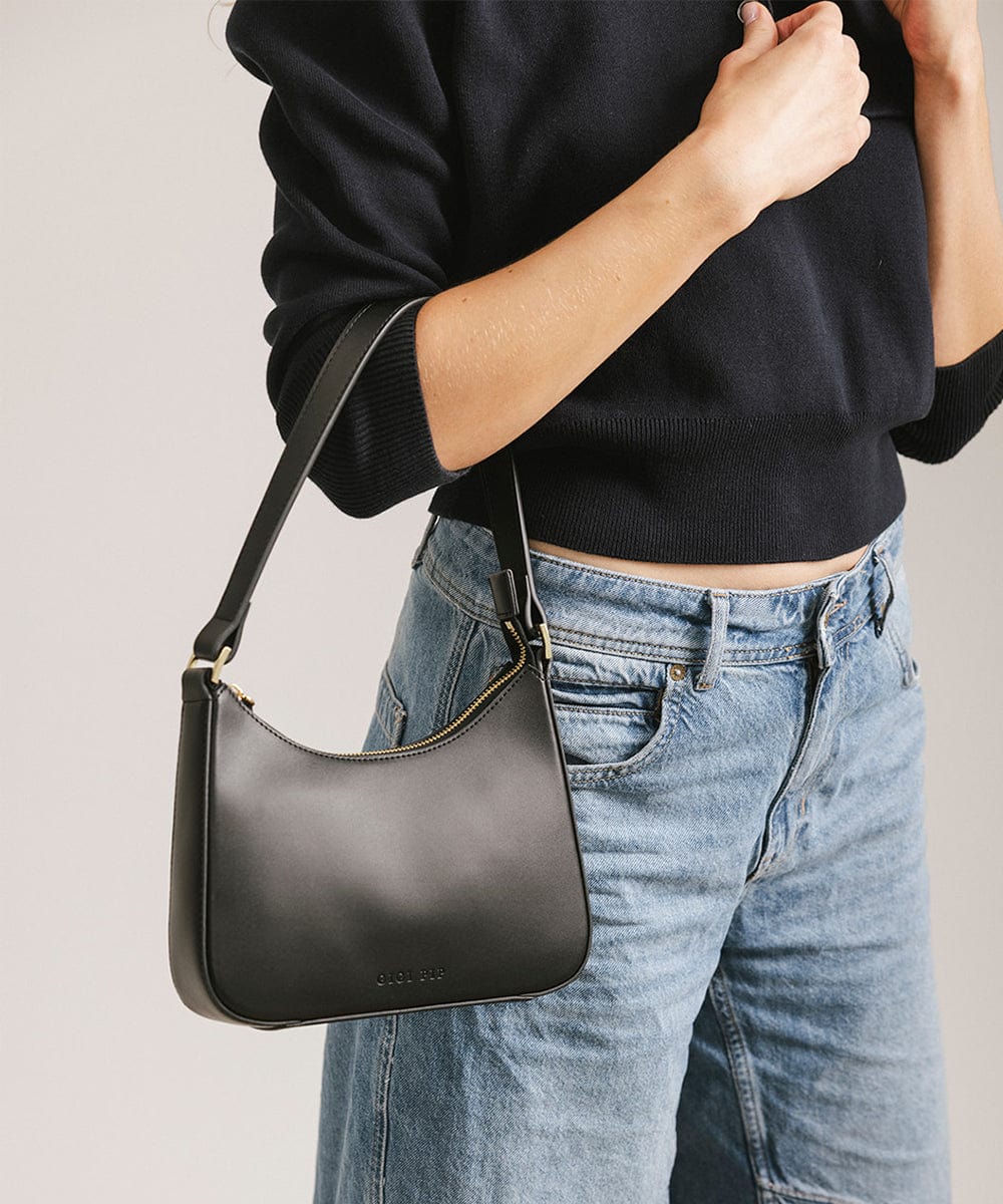 Person wearing a black shirt and blue jeans, carrying a black genuine leather handbag against a plain background #color_black