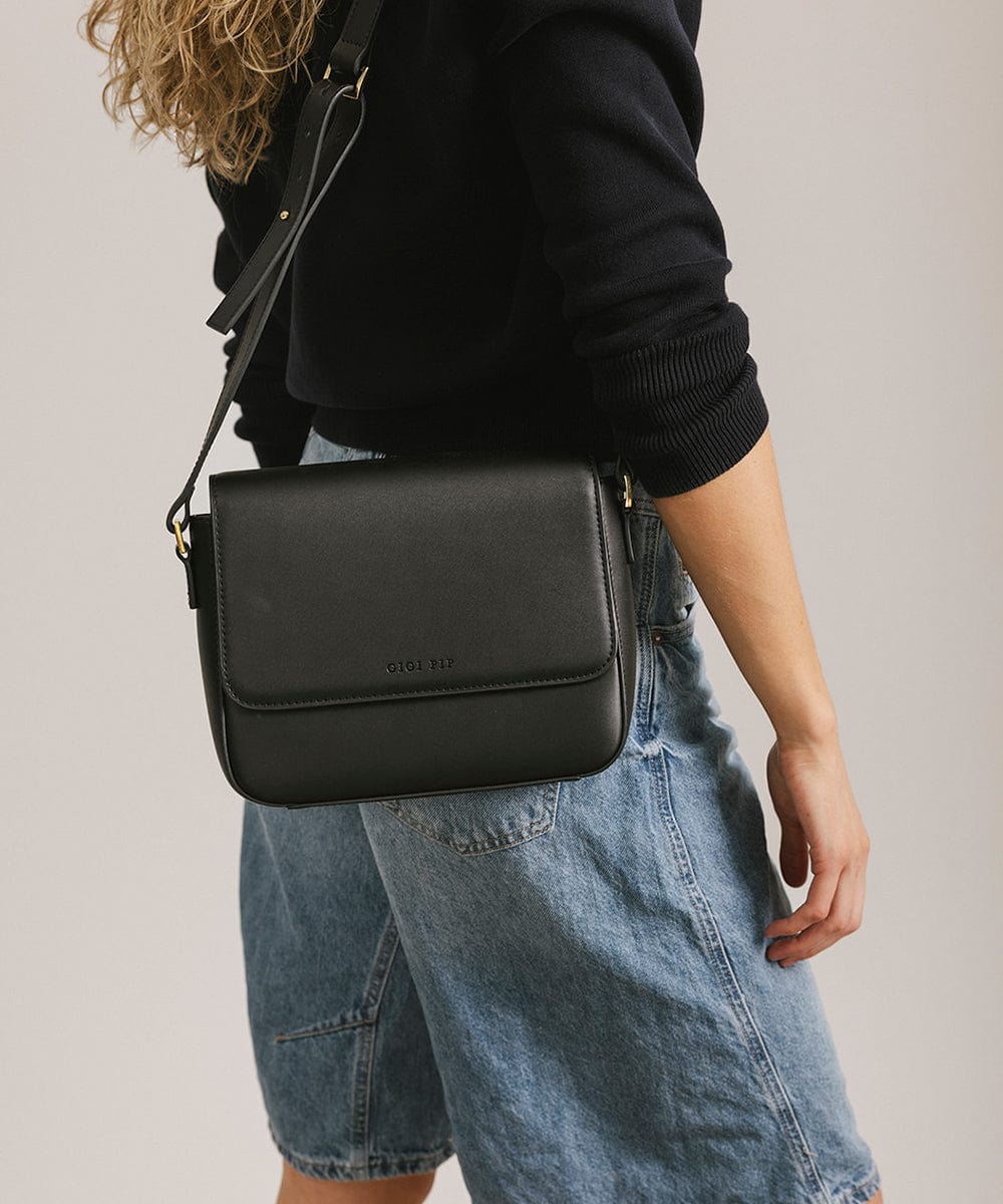 Person wearing a black sweater and denim shorts carrying a black Rhys Crossbody Genuine Leather Bag against a plain background #color_black