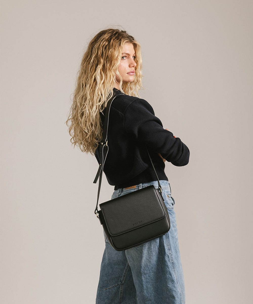 Woman wearing a black sweater and blue jeans carrying a black Rhys Crossbody Genuine Leather Bag, posing sideways against a plain background #color_black