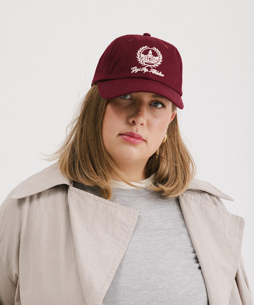 Woman wearing a burgundy ball cap with a white logo and text and a grey t-shirt with a beige shirt against a plain background #color_burgundy