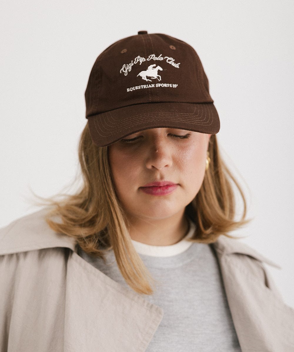 Woman wearing a brown cap with white text and horse logo and a grey t-shirt with a beige jacket against a plain background #color_chocolate