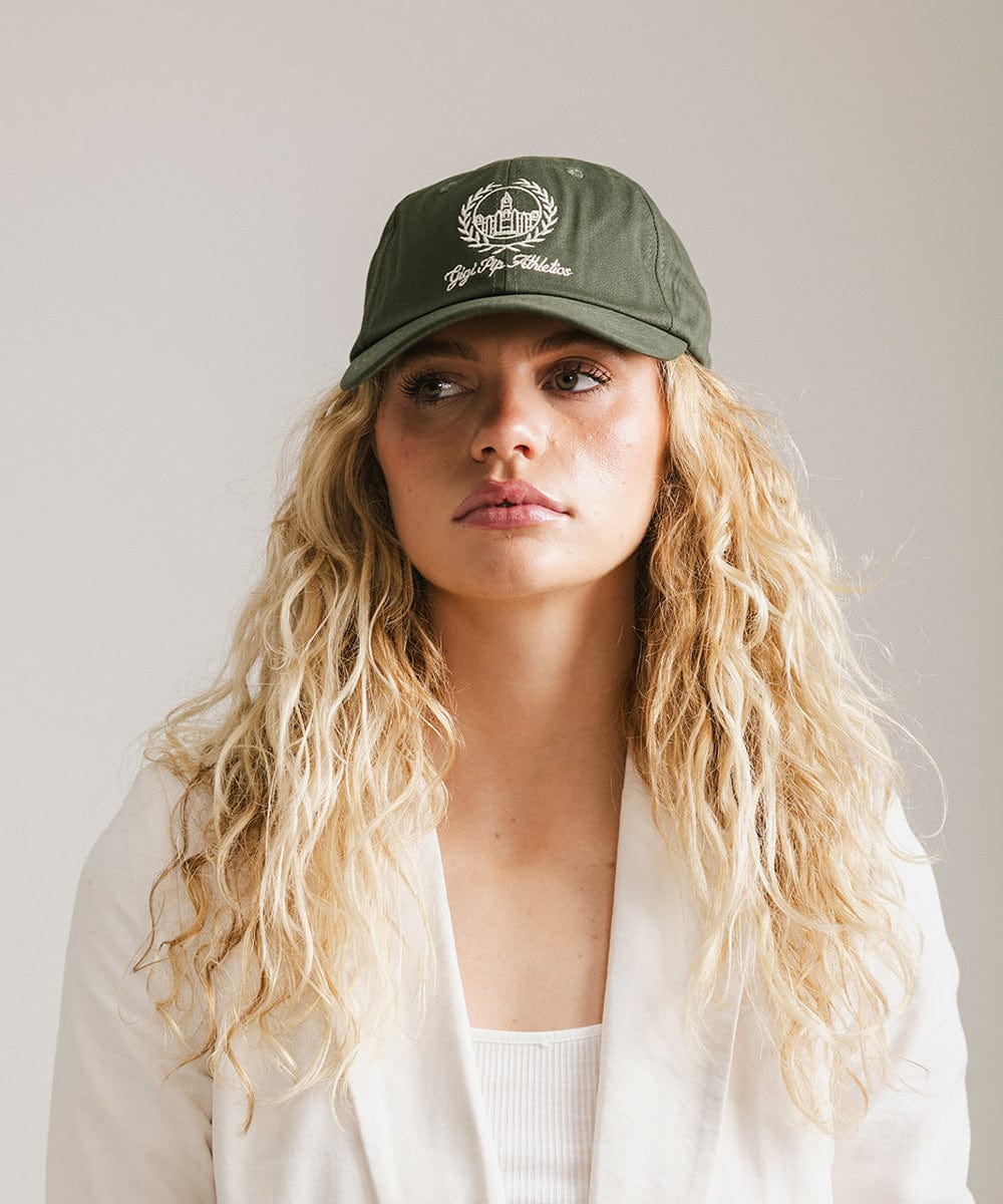 Woman wearing a dark green ball cap with a white logo and text and a white blazer against a plain background #color_dark green