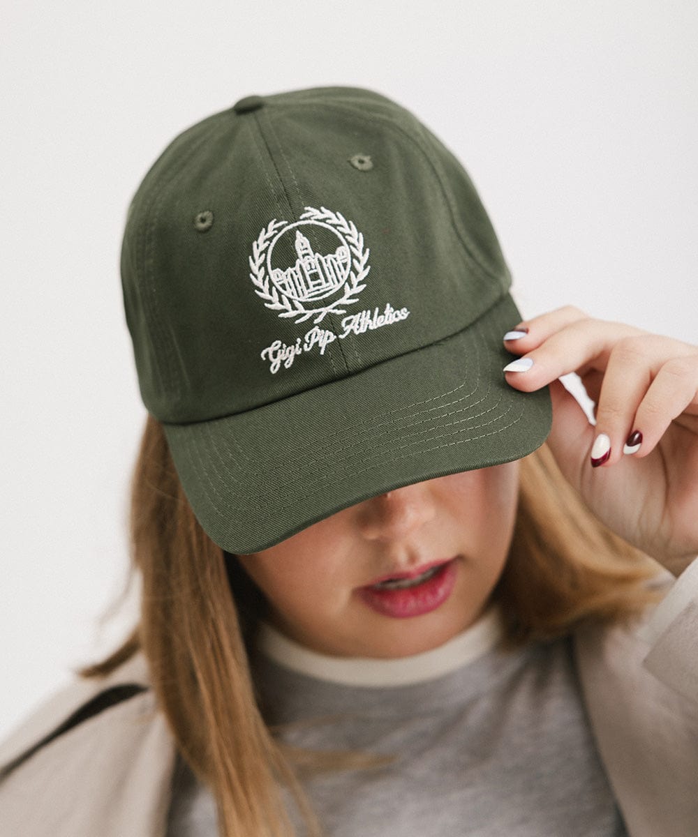 Woman wearing a dark green ball cap with a white logo and text and a grey shirt against a plain background #color_dark green