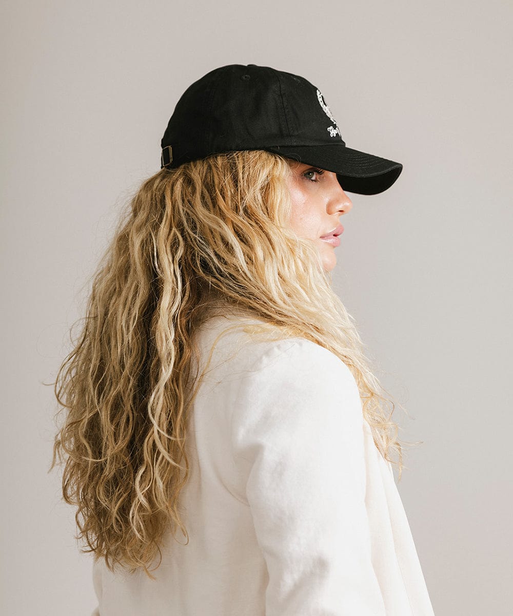 Woman wearing a black ball cap with a white logo and text and a white blazer, facing sideways against a plain background #color_black