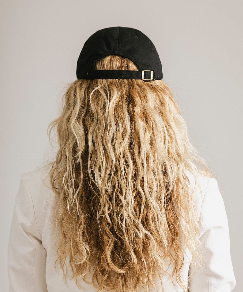 Woman wearing a black ball cap and a white blazer, facing behind against a plain background #color_black