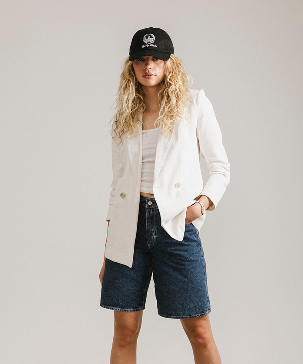 Woman wearing a black ball cap with a white logo and text, a white top with a white blazer and denim shorts against a plain background #color_black