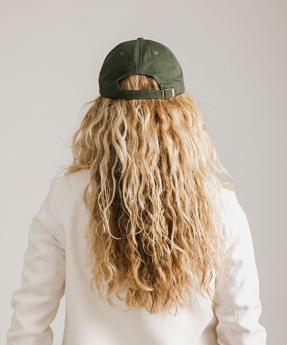 Woman wearing a dark green ball cap and a white blazer, facing behind against a plain background #color_dark green