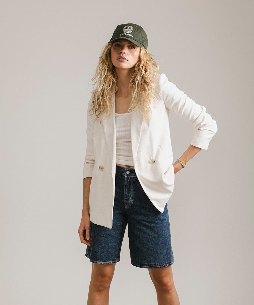 Woman wearing a dark green ball cap with a white logo and text, a white top with a white blazer and denim shorts against a plain background #color_dark green