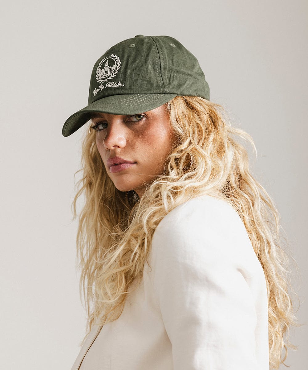 Woman wearing a dark green ball cap with a white logo and text and a white blazer, posing against a plain background #color_dark green