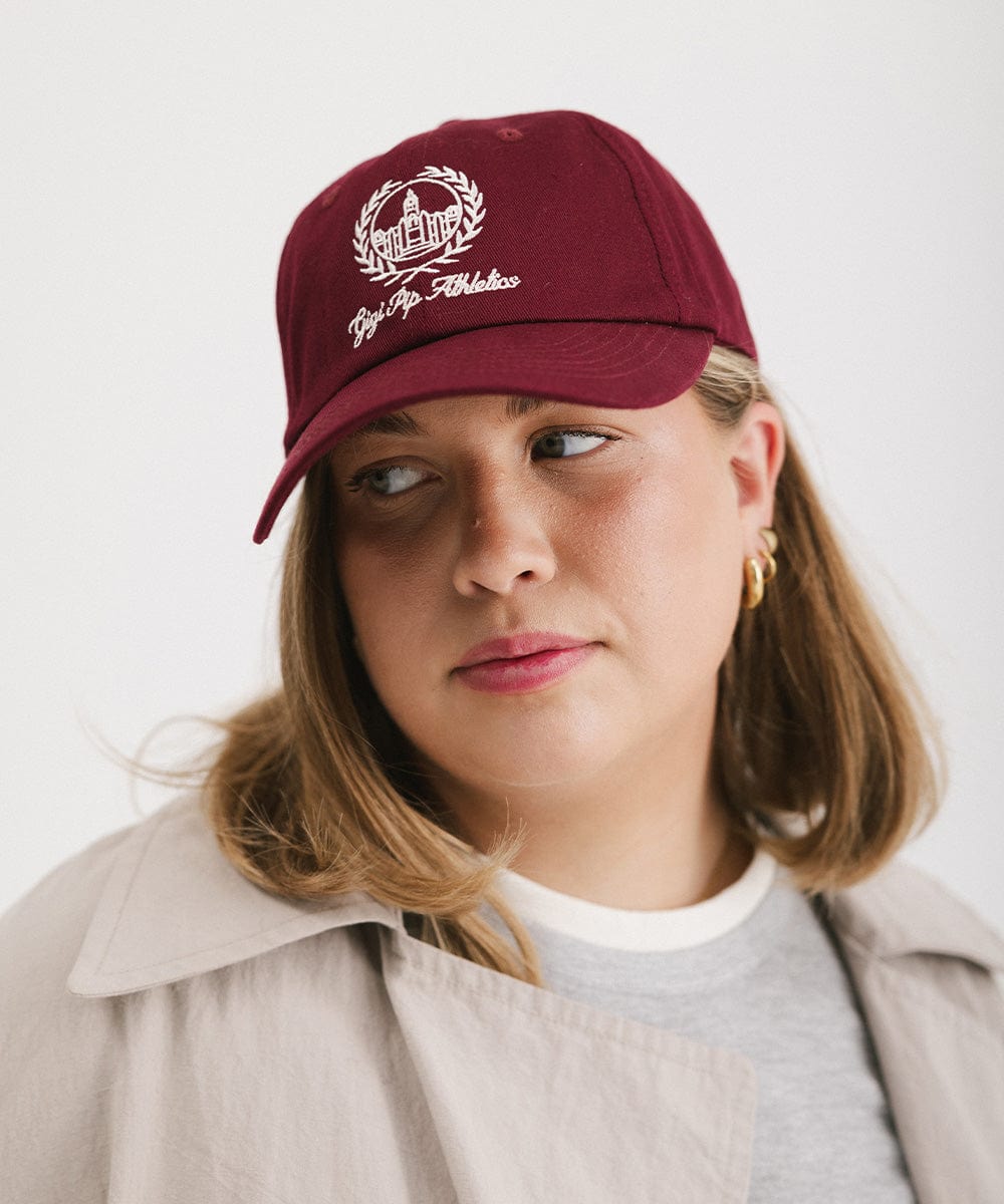 Woman wearing a burgundy ball cap with a white logo and text and a grey t-shirt with a beige shirt, looking sideways against a plain background #color_burgundy