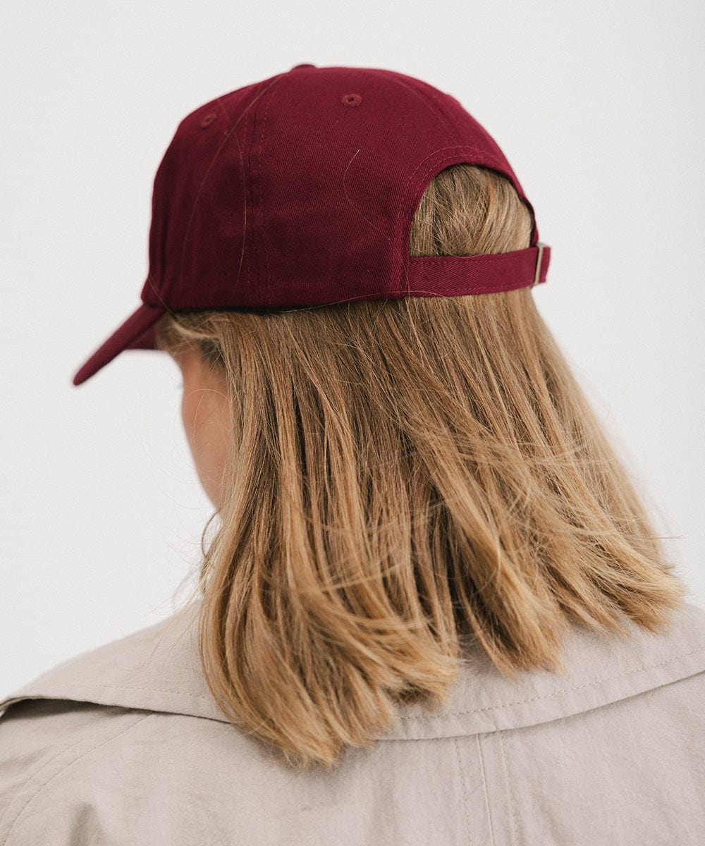Woman wearing a burgundy ball cap and a beige shirt, facing behind against a plain background #color_burgundy