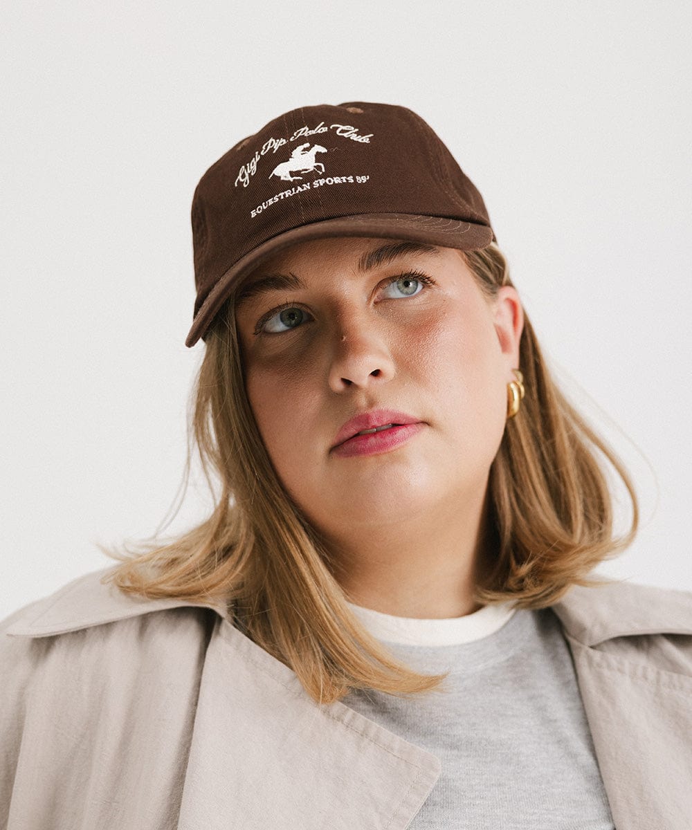 Woman wearing a brown cap with white text and horse logo and a grey t-shirt with a beige jacket against a plain background #color_chocolate