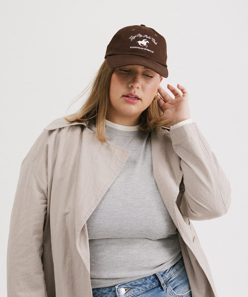 Woman wearing a brown cap with white text and horse logo and a grey t-shirt with a beige jacket against a plain background #color_chocolate