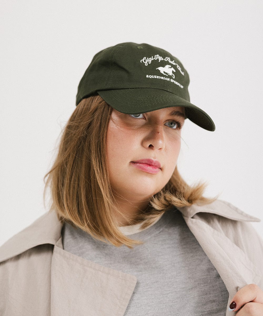 Woman wearing a dark green cap with white text and horse logo and a grey t-shirt with a beige jacket, posing against a plain background #color_dark green