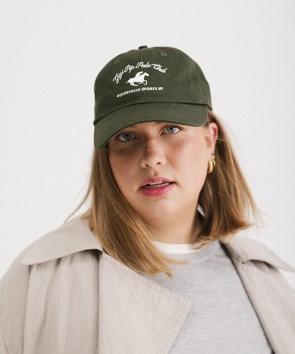 Woman wearing a dark green cap with white text and horse logo and a grey t-shirt with a beige jacket against a plain background #color_dark green