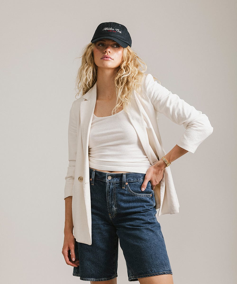 Woman with long hair wearing a navy cap with white 'Athletics Club' text, a white top with a white shirt and denim shorts against a plain background #color_navy