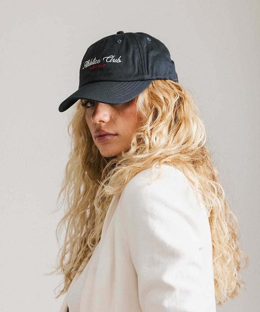 Woman with long hair wearing a navy cap with white 'Athletics Club' text and a white shirt, posing sideways against a plain background #color_navy