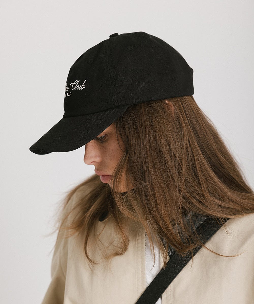 Woman wearing a black cap with white 'Athletics Club' text and a beige jacket, facing sideways against a plain background #color_black