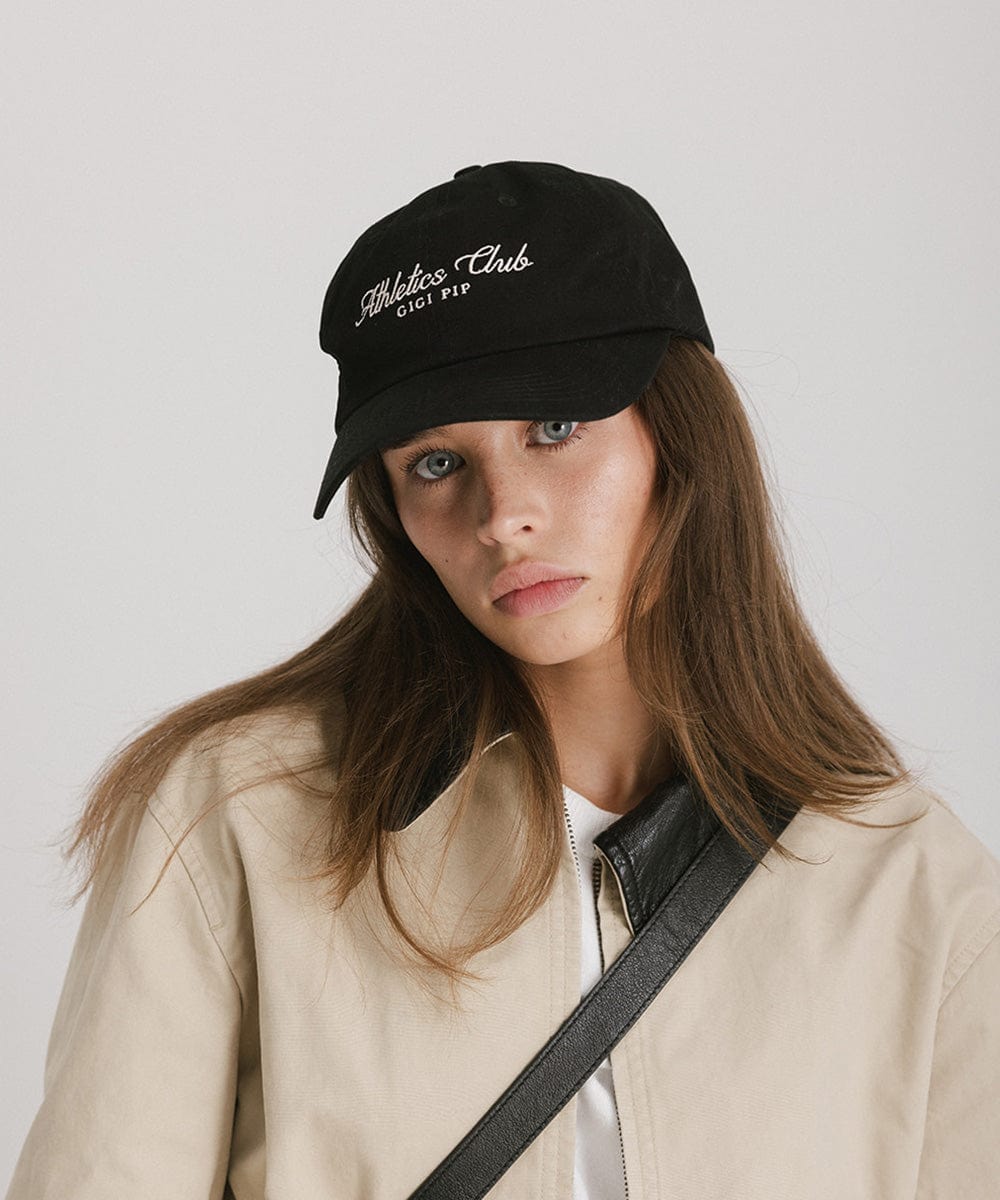 Woman wearing a black cap with white 'Athletics Club' text and a beige jacket against a plain background #color_black