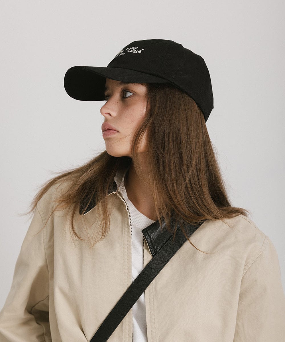 Woman wearing a black cap with white 'Athletics Club' text and a beige jacket, posing sideways against a plain background #color_black