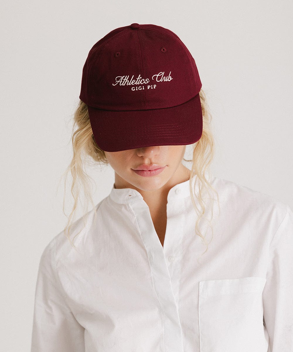 Woman wearing a burgundy cap with white 'Athletics Club' text and a white shirt against a plain background #color_burgundy