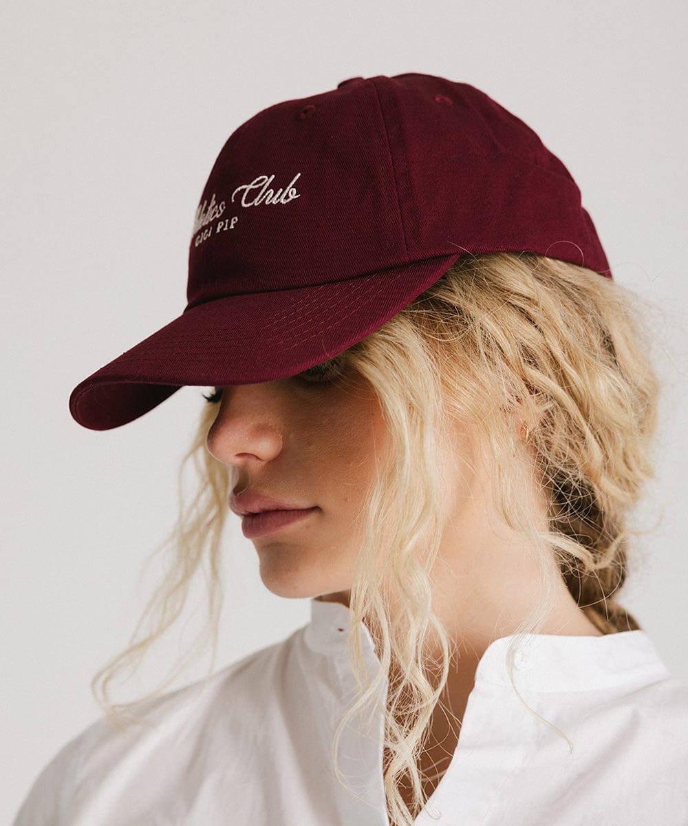 Woman wearing a burgundy cap with white 'Athletics Club' text and a white shirt, posing sideways against a plain background #color_burgundy