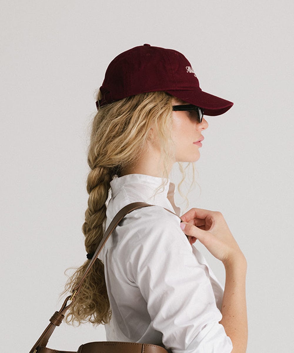 Woman wearing a burgundy cap, sunglasses and a white shirt, facing sideways against a plain background #color_burgundy