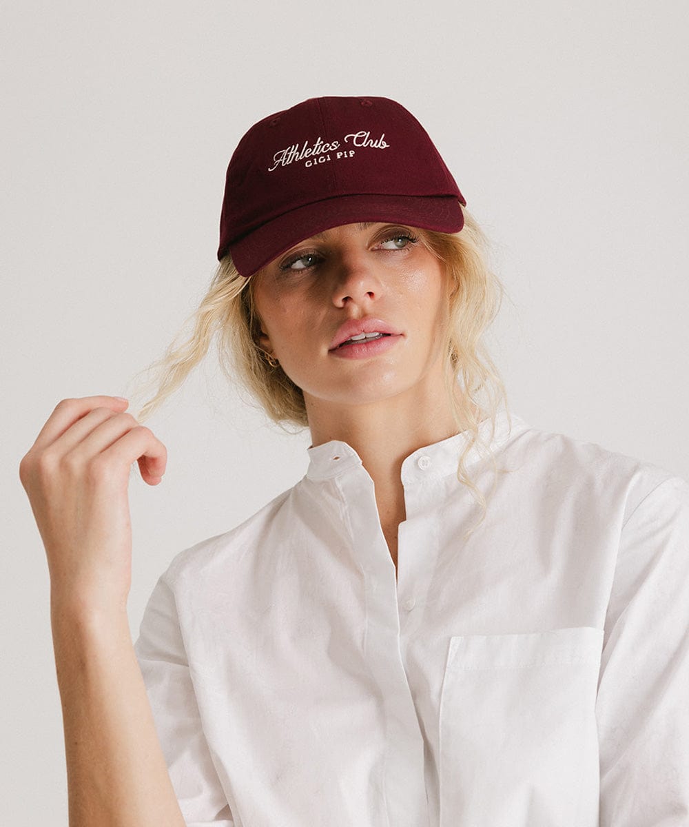 Woman wearing a burgundy cap with white 'Athletics Club' text and a white shirt against a plain background #color_burgundy