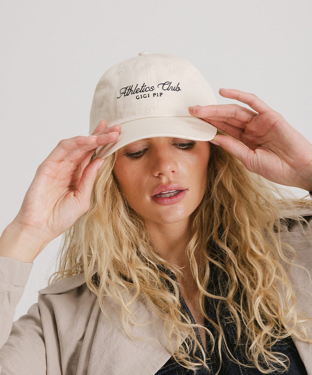 Woman wearing a cream cap with black 'Athletics Club' text and a denim top with a beige shirt against a plain background #color_cream