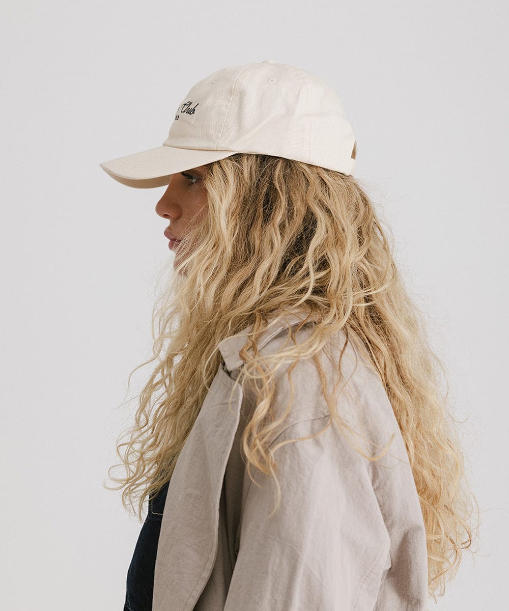 Woman wearing a cream cap with black 'Athletics Club' text and a beige shirt, facing sideways against a plain background #color_cream