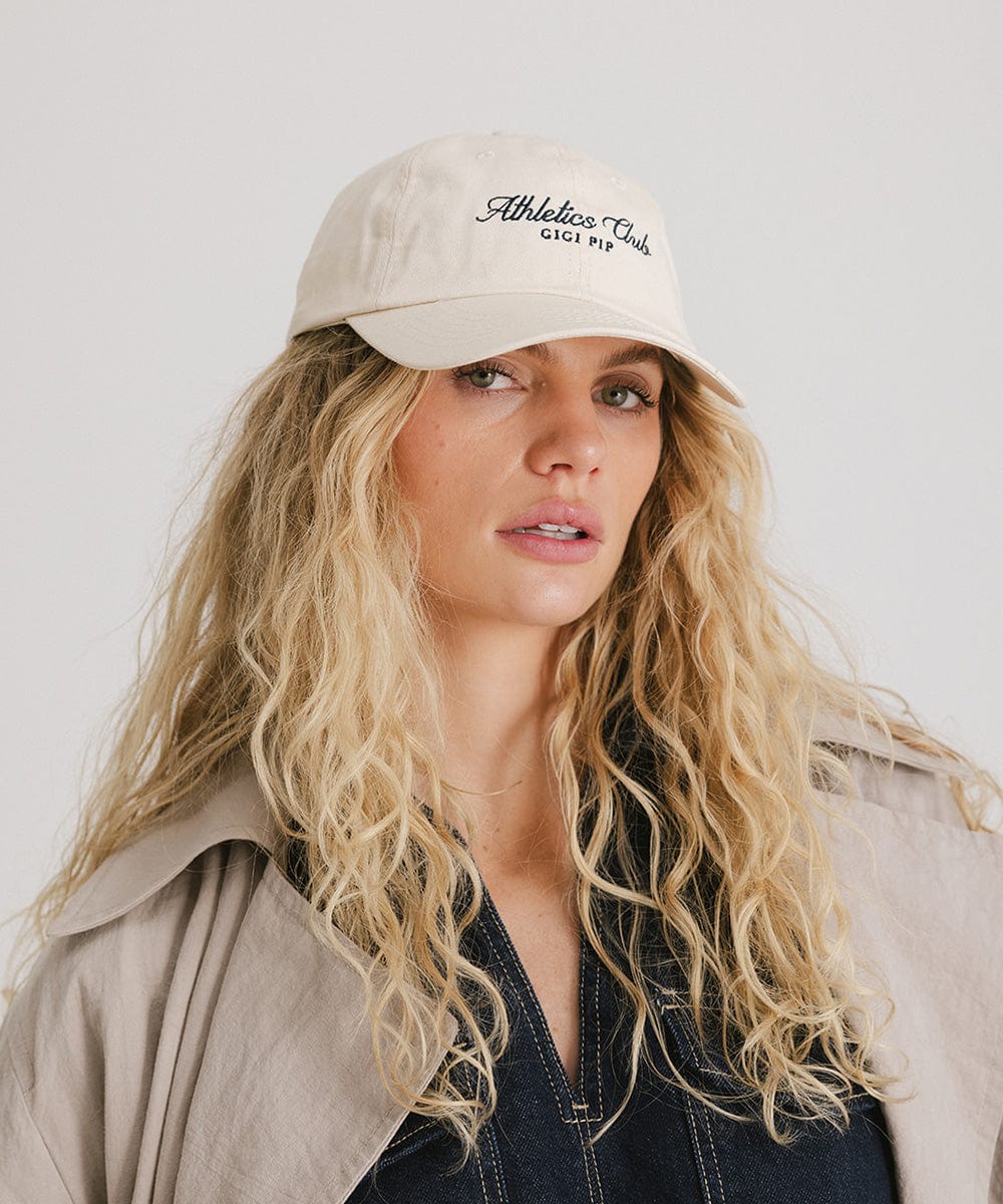 Woman wearing a cream cap with black 'Athletics Club' text and a denim top with a beige shirt against a plain background #color_cream
