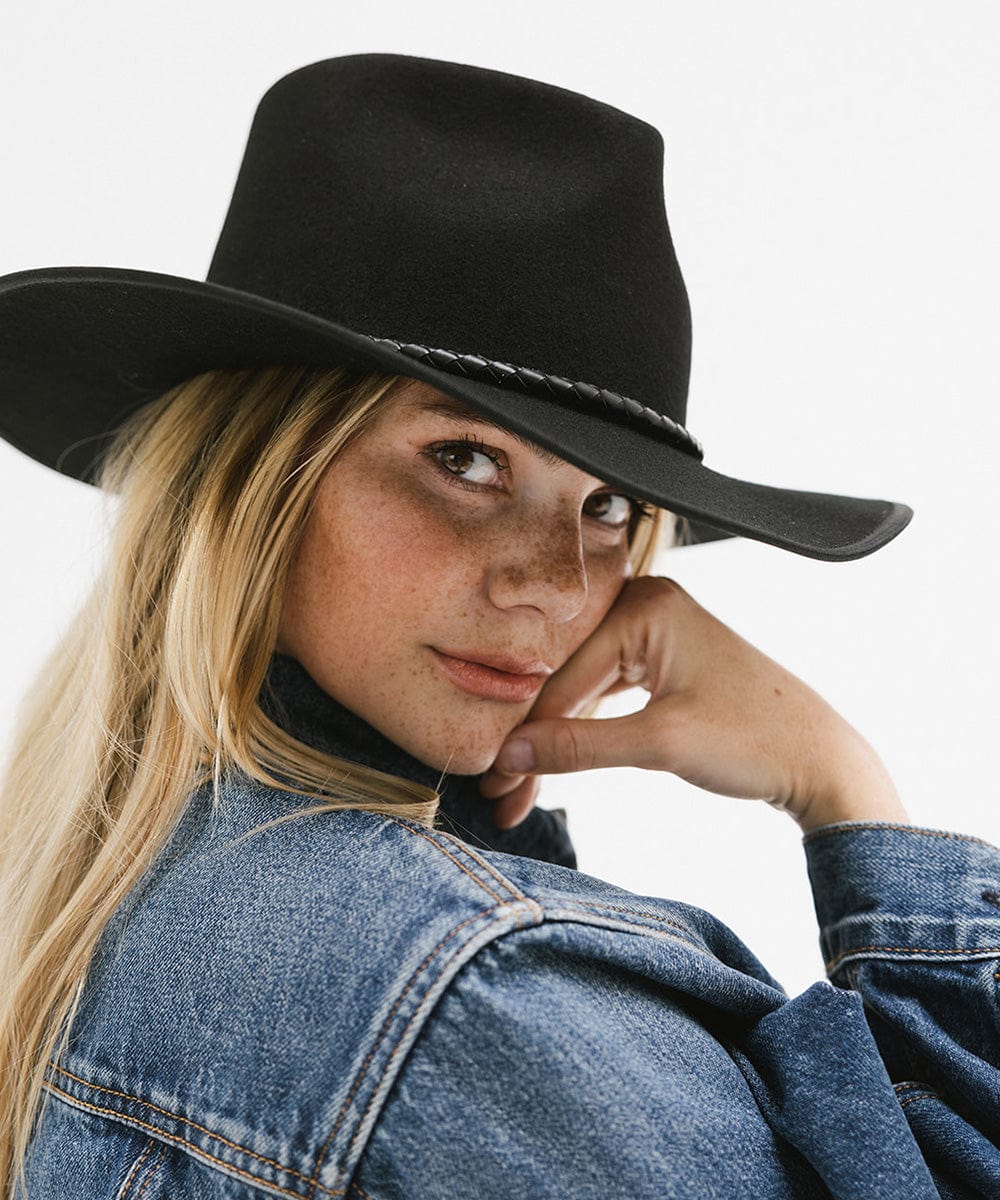 Woman wearing a black hat with a black braided bolo hat band and a denim jacket, posing sideways against a plain background #color_black