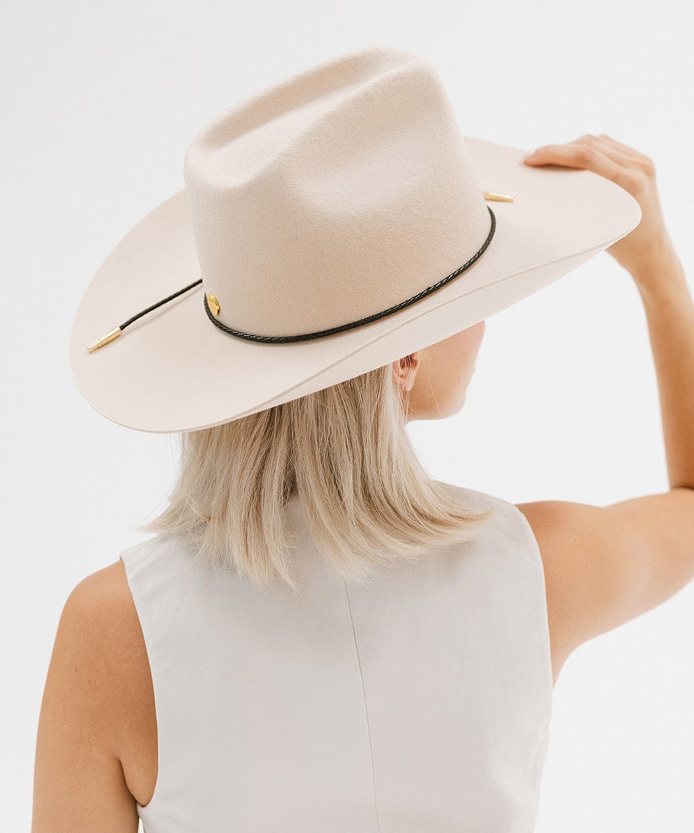 Woman wearing a cream cowboy hat with a black braided hat band and a white top, facing behind against a plain background #color_black