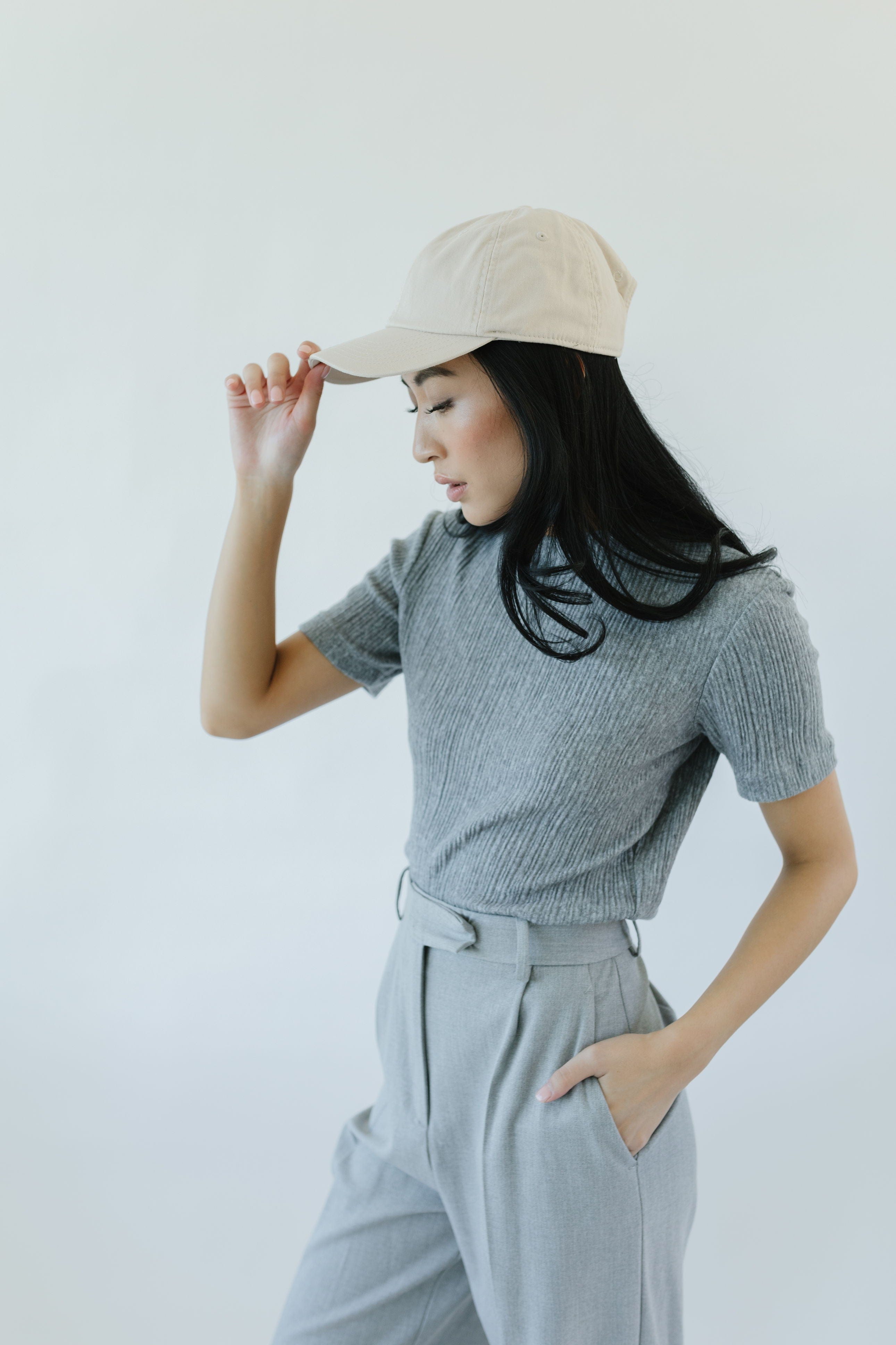 Woman wearing a cream colored ball cap, a grey t-shirt and grey pants, facing sideways against a plain background #color_cream
