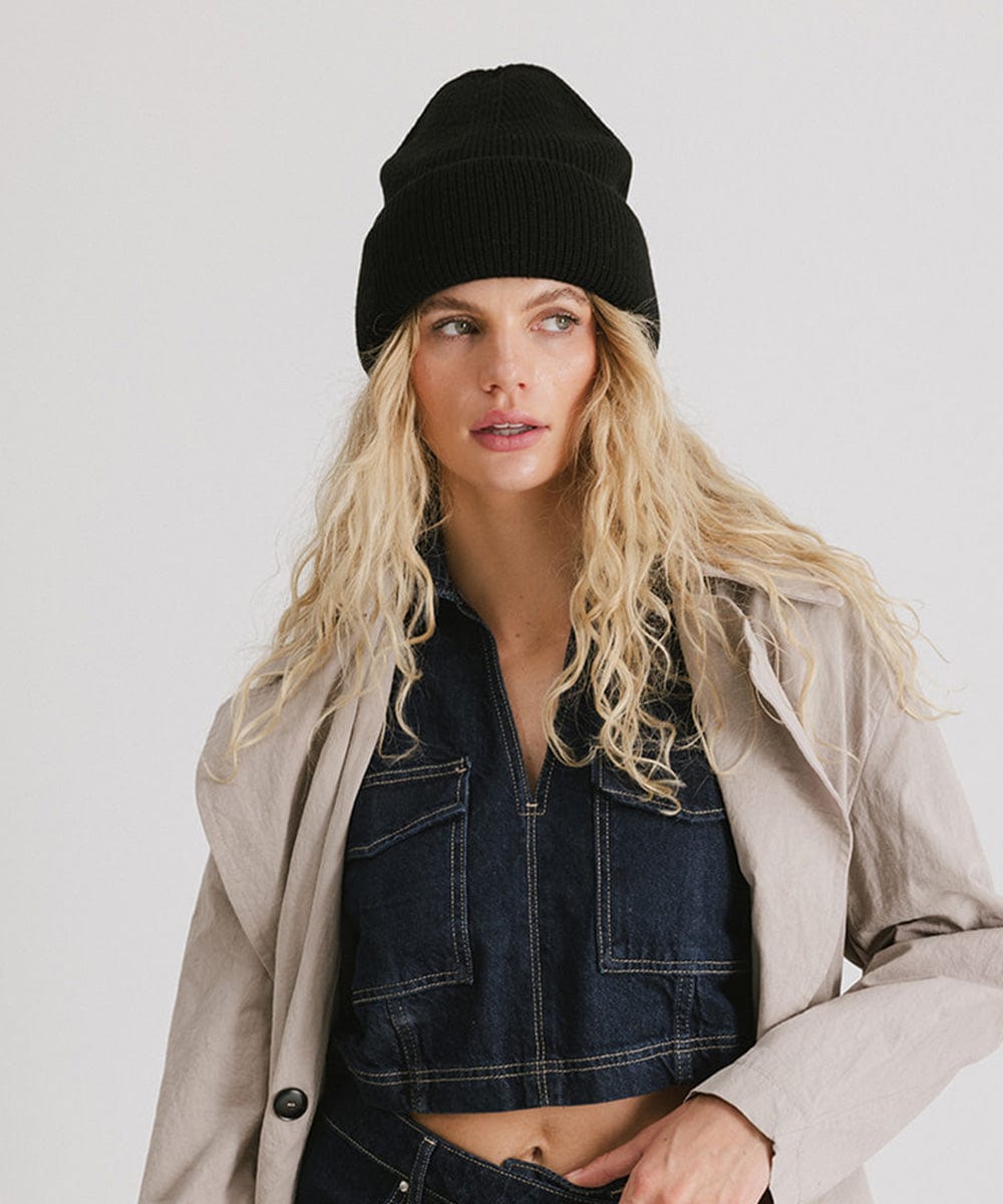 Woman with blonde hair wearing a black oversized knit beanie and a denim top with a cream coat against a plain background #color_black