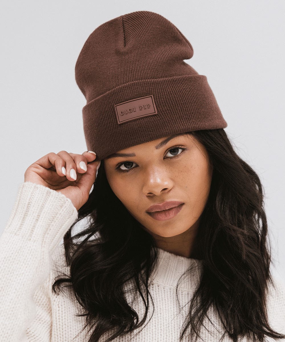 Woman wearing a chocolate colored knit beanie and a white sweater against a plain background #color_chocolate