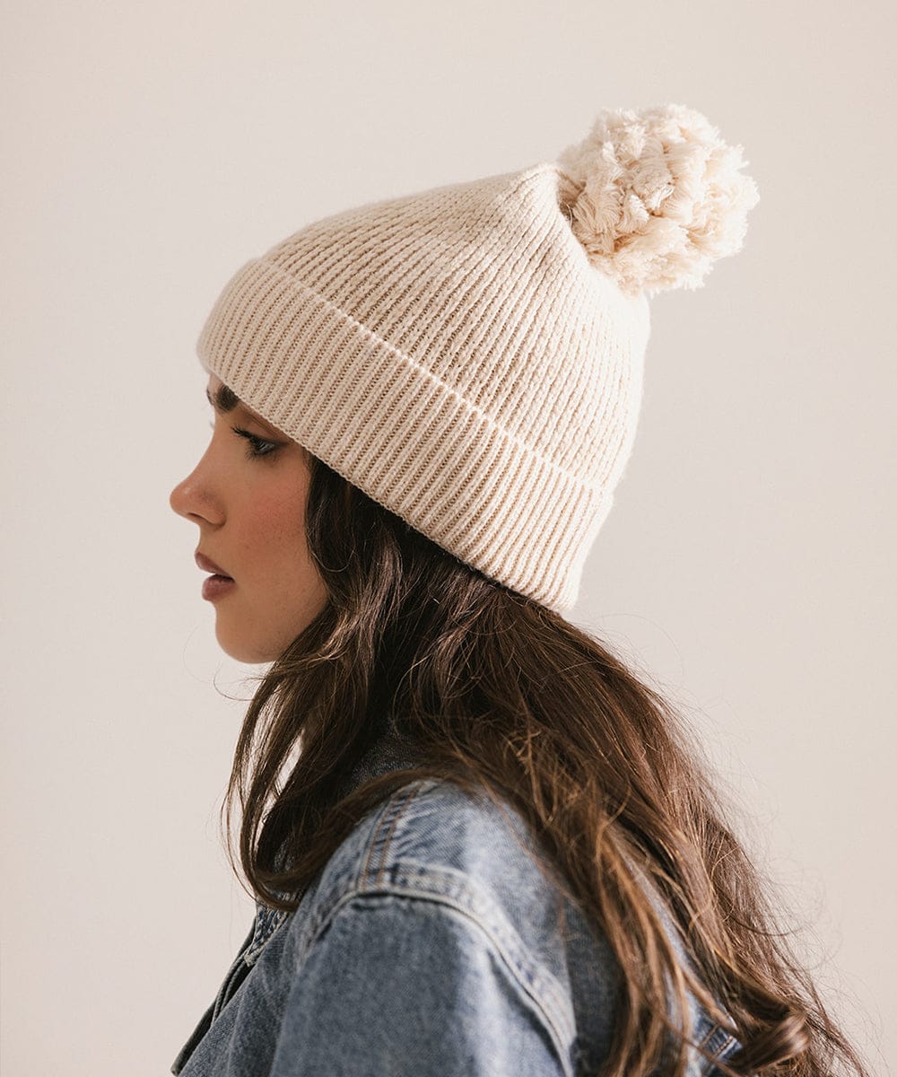 Woman wearing a beige knit beanie with a white pom-pom against a plain background #color_cream